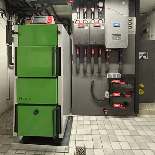 Großer grüner Luuma Holzvergaser links. Im Hintergrund Installationskomponenten an grauer Wand des Pufferspeichers installiert